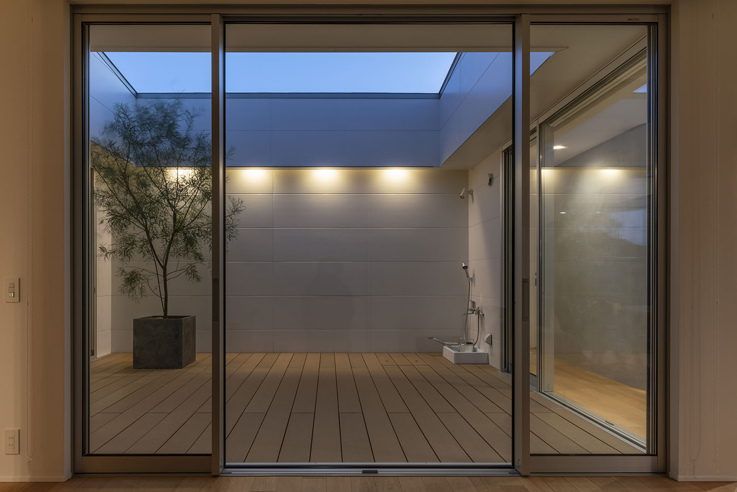 シンボルツリーがあるウッドデッキの中庭の壁に、照明が取り付けられた様子・デザイン住宅 注文住宅・デザイン住宅建築実例_『CASE661 環(めぐる)いえ』_中庭