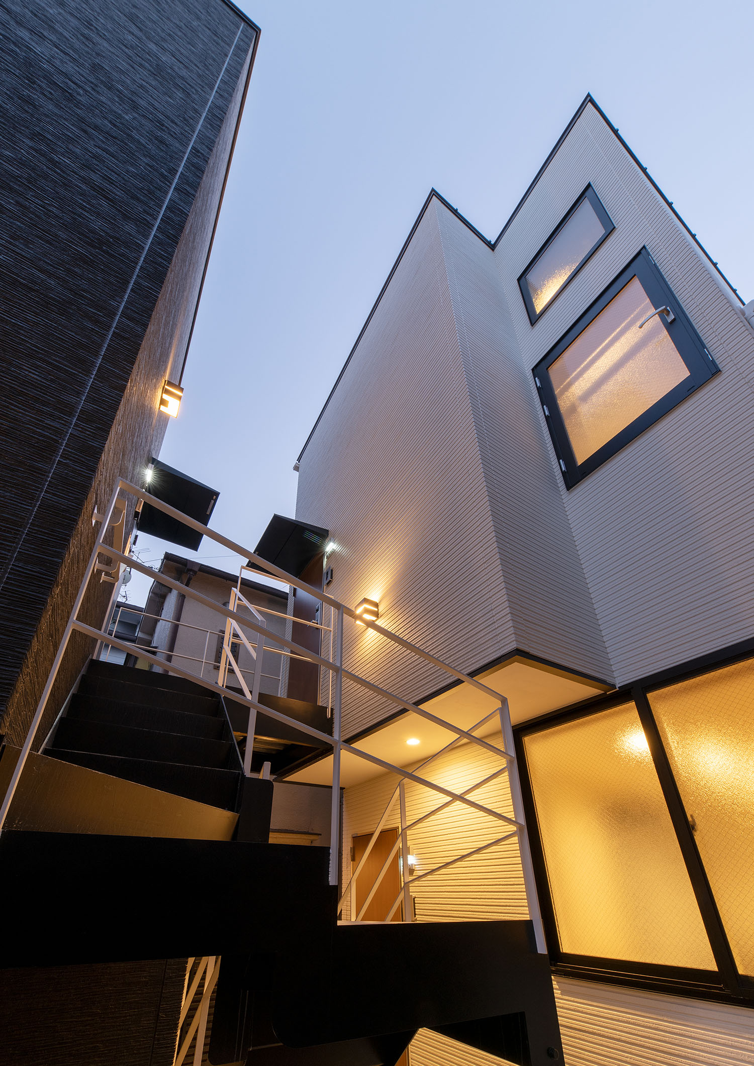 外壁に取り付けられた照明と、窓からの光で照らされる白い外壁の建物・賃貸併用住宅 注文住宅・デザイン住宅建築実例_『CASE681  penche'　「パンシェ」の家』_外観