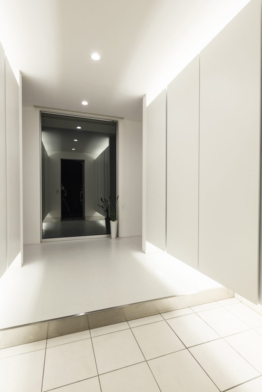 間接照明が取り付けられた浮いたデザインのシューズボックスがある、正面がガラス窓の玄関。