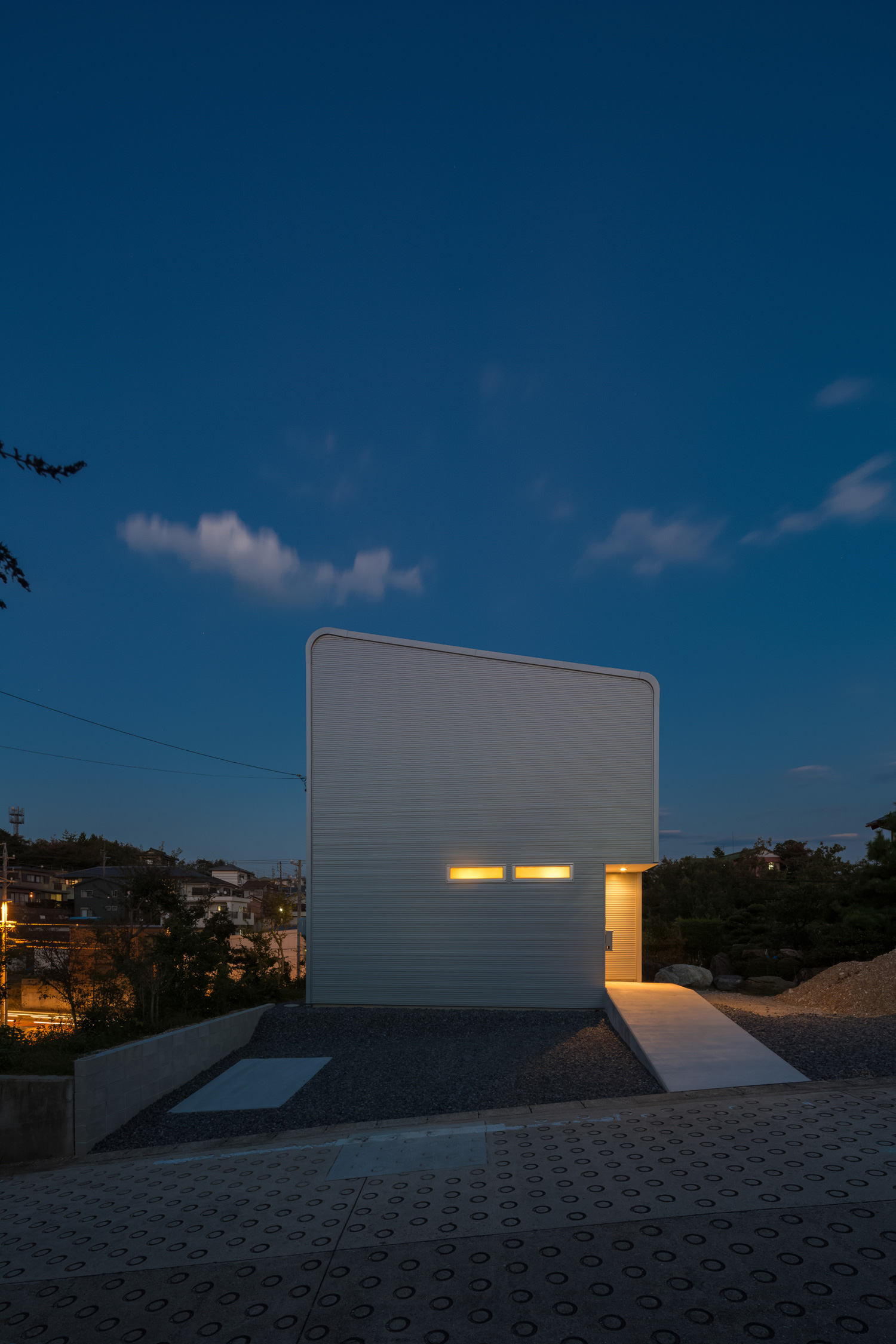 コンクリートのアプローチの先にある照明に照らされた玄関・デザイン住宅