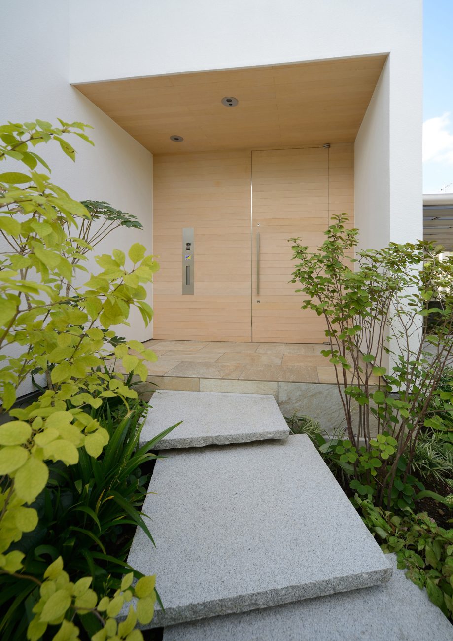 明るい色の木材の軒天井と玄関扉がある植物に囲まれた玄関・デザイン住宅