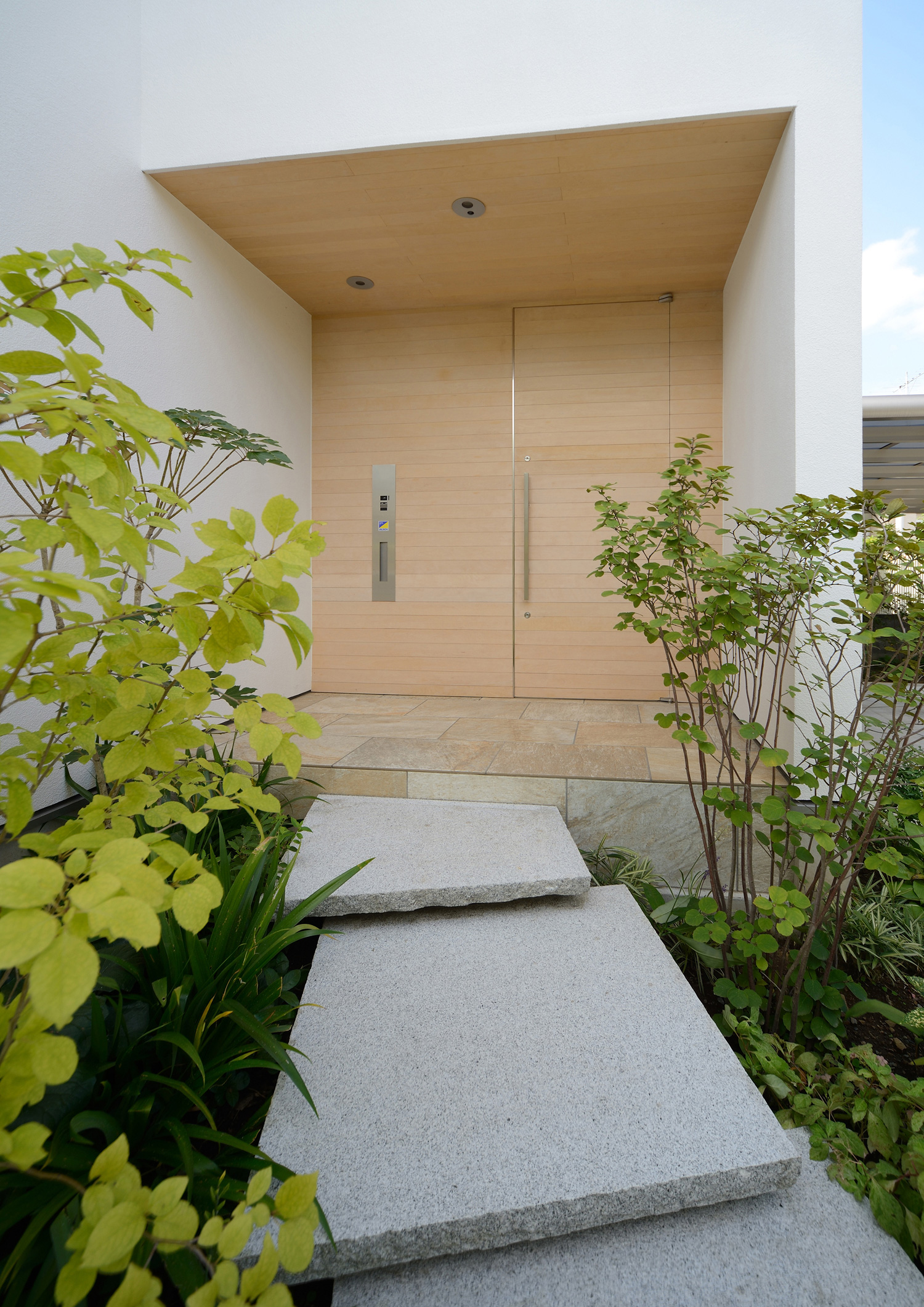 明るい色の木材の軒天井と玄関扉がある植物に囲まれた玄関・デザイン住宅