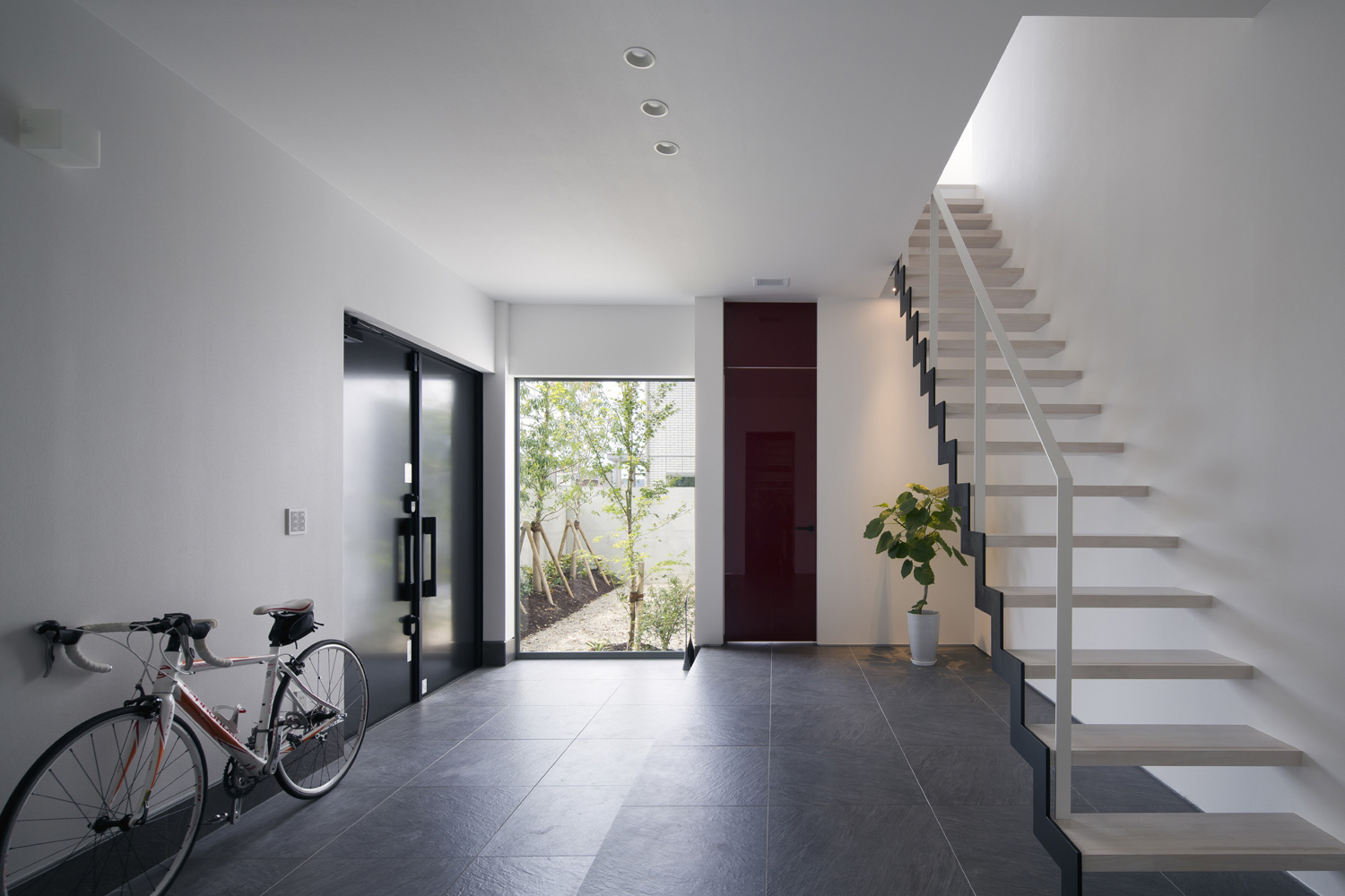 大きな窓から植栽が見える玄関とスケルトン階段のある廊下・デザイン住宅