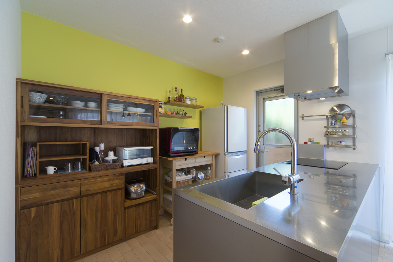 ステンレスのペニンシュラキッチンの背面にある木のカップボードとアクセントクロス・デザイン住宅