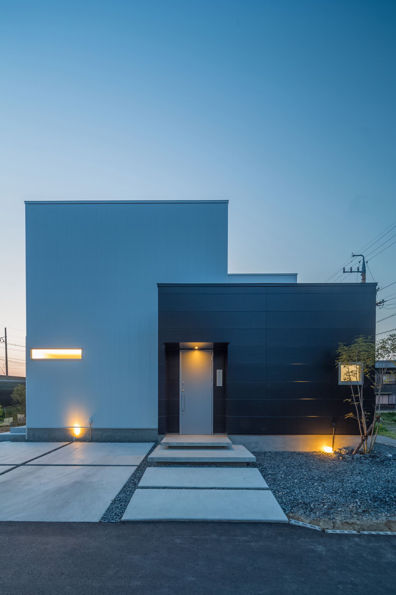 照明にライトアップされたコンクリートのアプローチがあるモノトーンの外壁の住まい・デザイン住宅