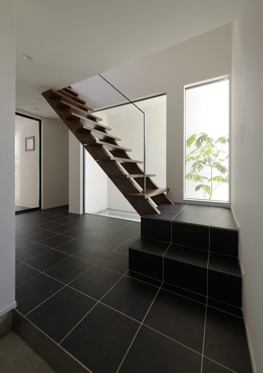 正面に植栽が見える窓やスケルトン階段がある黒いタイルの玄関・デザイン住宅 注文住宅・デザイン住宅建築実例_『CASE514 Slanting Hat』_玄関