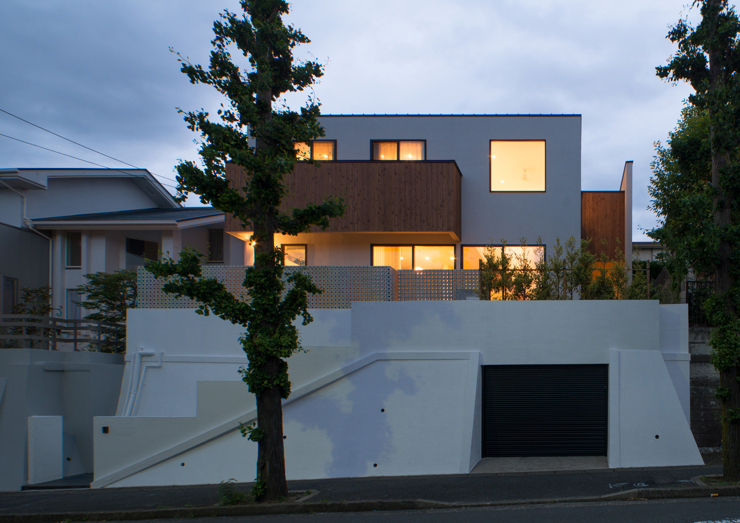 窓から光が漏れる、高台の上にあり前に木が植えられた家・デザイン住宅 注文住宅・デザイン住宅建築実例_『CASE530 ミクストメディア』_外観