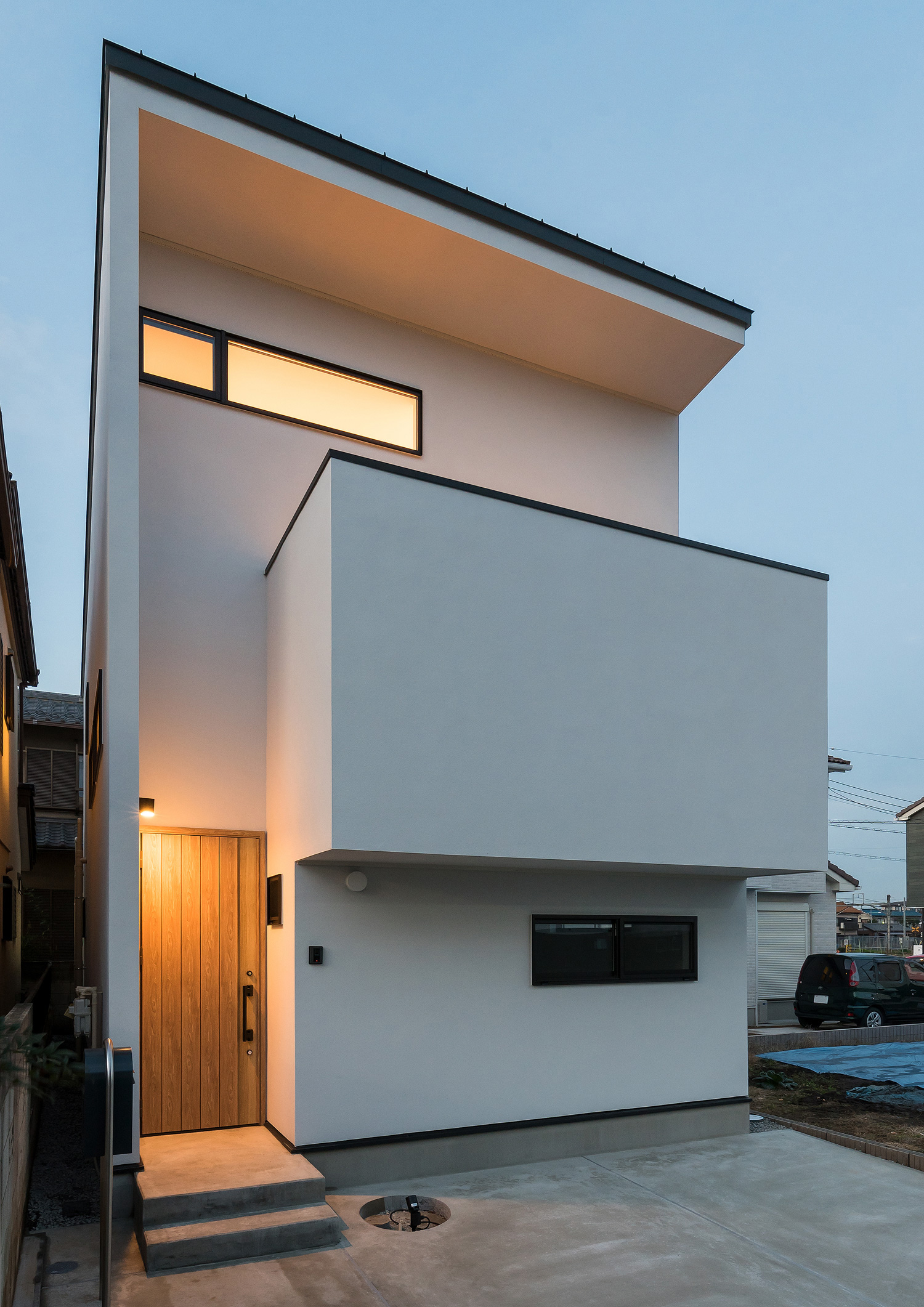 アプローチの壁に取り付けられた照明が木の玄関扉を照らす白い外壁の家・デザイン住宅 注文住宅・デザイン住宅建築実例_『CASE590 つなぐ家』_外観