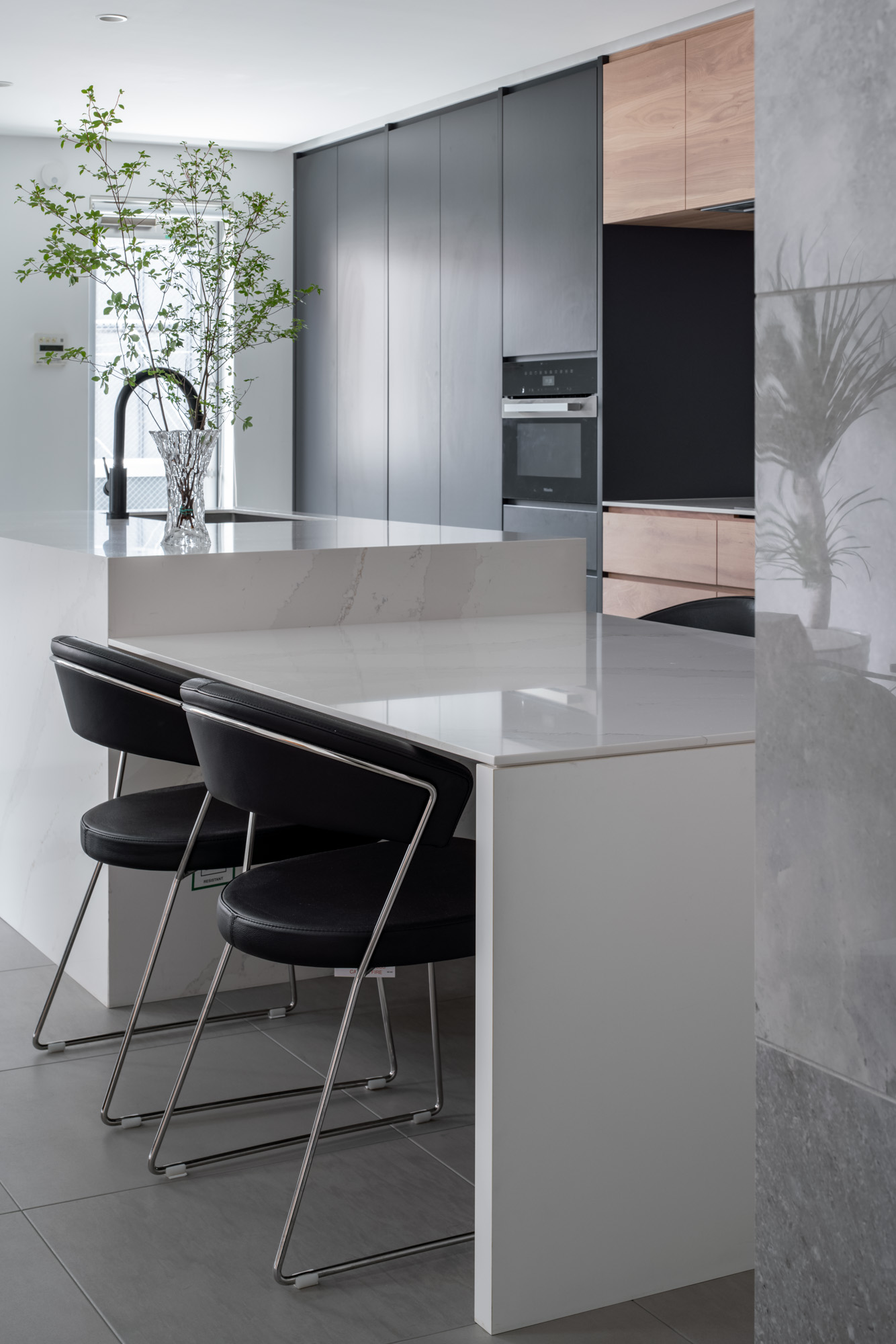 大理石風のキッチンに植物が飾られ、ダイニングテーブルには黒い椅子が置かれている様子・デザイン住宅 注文住宅・デザイン住宅建築実例_『CASE749 dignified housing』_LDK
