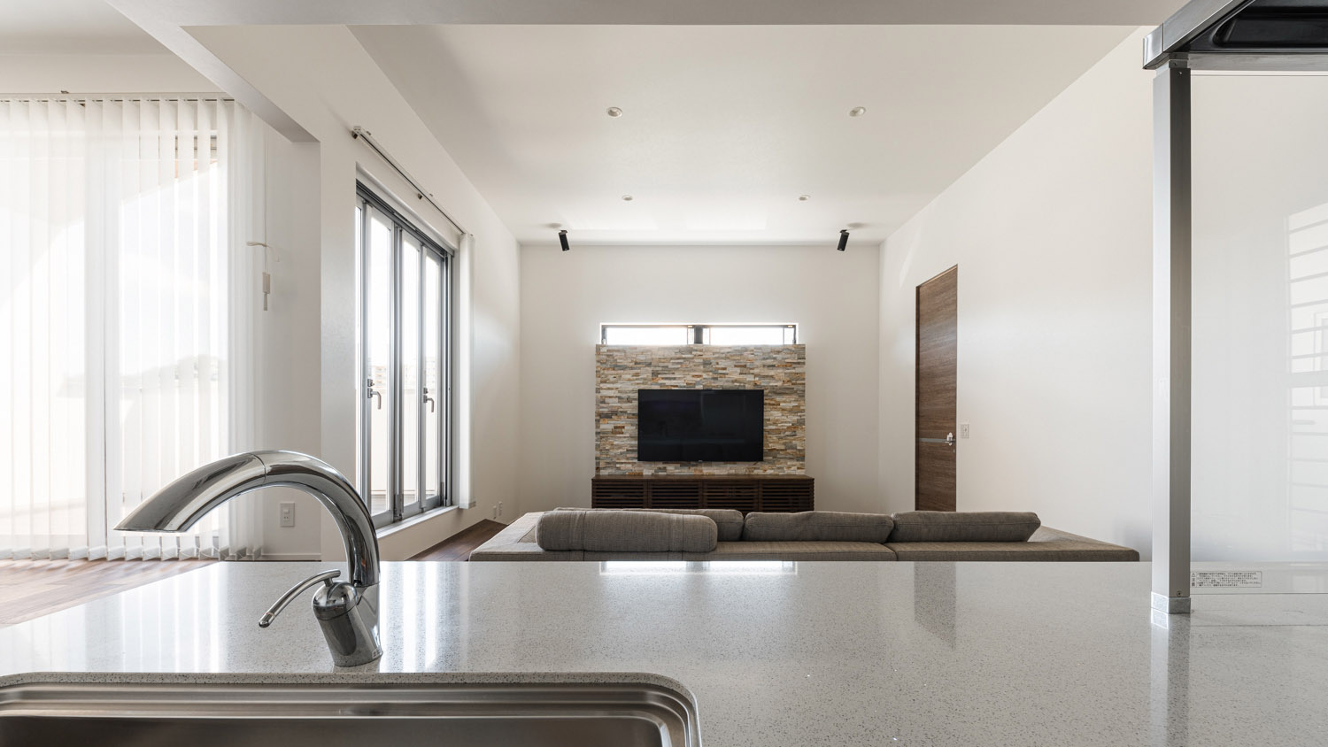 白いキッチンから、ソファーやアクセントウォールにかけられたテレビが見える様子・デザイン住宅 注文住宅・デザイン住宅建築実例_『CASE754 首途(かどで)の家』_LDK